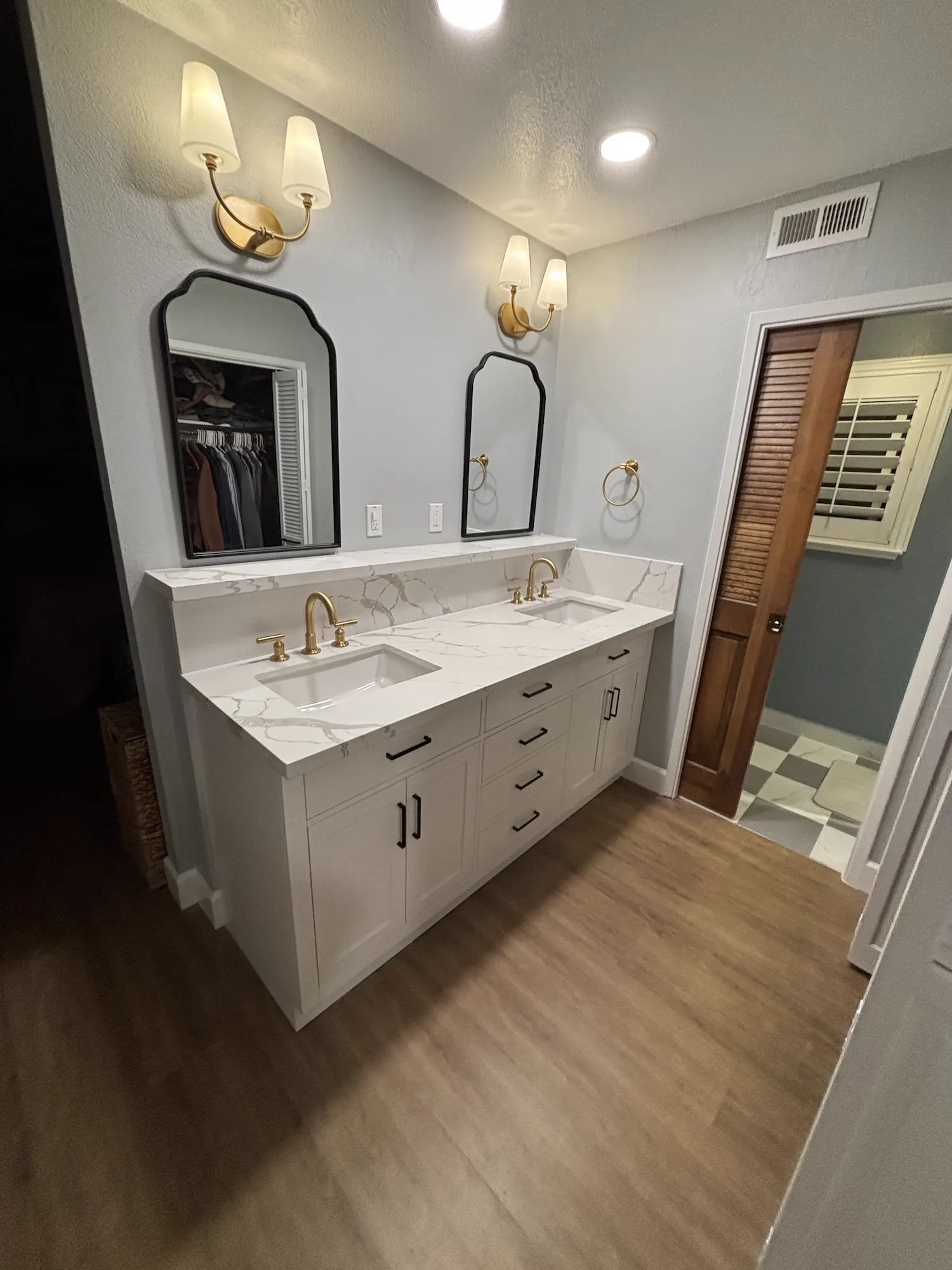 After: Master bath vanity rebuild: dated tile-top double vanity replaced with a white shaker vanity, quartz counter and backsplash, gold fixtures, and arched mirrors.